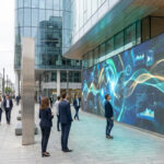 Grand écran numérique vibrant affichant des données abstraites dans une rue urbaine animée. Des passants en costume regardent l'écran ou marchent.