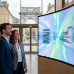 Deux professionnels regardent un grand écran LED incurvé diffusant des graphiques lumineux dans un hall moderne avec vue sur la ville.