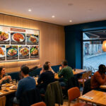 Grand écran digital lumineux dans un restaurant moderne, affichant des plats avec prix, entouré de clients attablés.